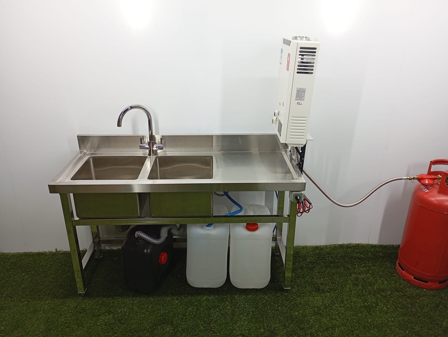 Stainless steel sink with a water heater and gas canister on a grassy floor.